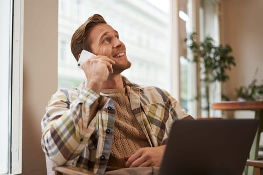 Portrait of young businessman, freelancer working remotely from coffee shop in city, sitting in chair with laptop, answer a phone call, talking on mobile to someone.