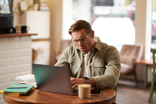 Portrait of angry man in glasses, looking frustrated and typing something on laptop, staring at screen with outraged face expression, sitting in cafe.