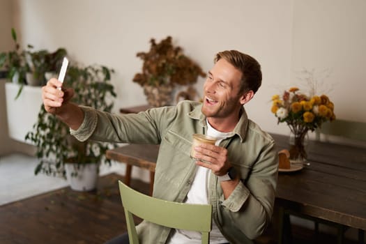 Portrait of handsome happy young man, taking selfie in cafe, posing with cup of coffee, video chats using smartphone app, talks to a friend. Lifestyle and leisure concept
