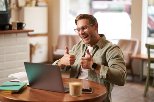 Portrait of young smiling man, online tutor shows thumbs up at laptop, video chats with someone who did good job, guy expresses approval.