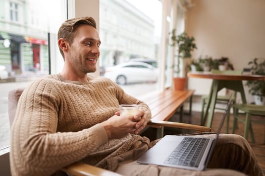 Portrait of handsome young man sits in cafe, drinks coffee and watches video on laptop, looking at screen with happy smile, relaxing in co-working space.