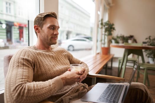 Portrait of male entrepreneur, digital nomad sitting in cafe with glass of cappuccino, looking at laptop, watching video, attending meeting online from coffee shop.
