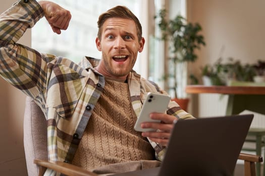 Portrait of funny athletic, fit guy in cafe, sitting on chair with laptop and smartphone, flexing biceps, showing his muscles on camera and bragging how strong he is.