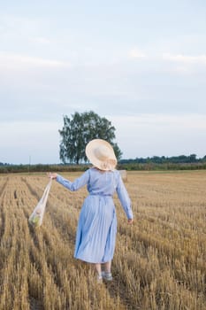 A red-haired woman in a hat and a blue dress walks in a field with haystacks. The view from the back