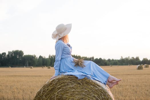 A red-haired woman in a hat and a blue dress walks in a field with haystacks