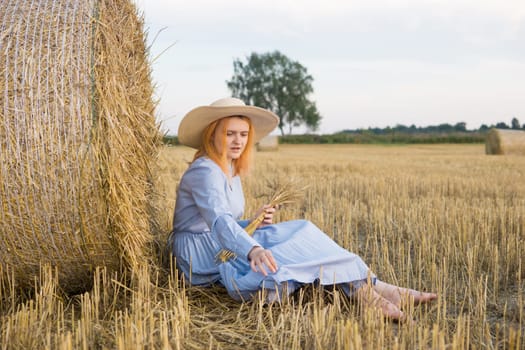 A red-haired woman in a hat and a blue dress walks in a field with haystacks