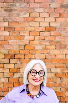 portrait of a beautiful senior woman with glasses smiling happy looking at camera with a brick wall in the background, concept of happiness of elderly people and active lifestyle, copy space for text