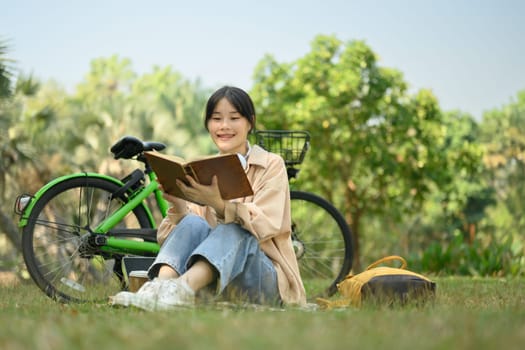 Carefree young woman reading book on green grass at the park with a bicycle.