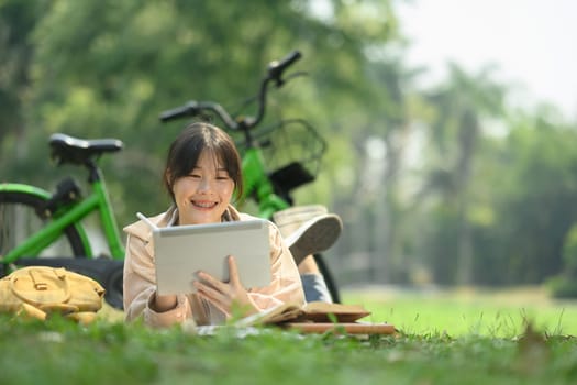 Happy young woman lying on green grass near her bicycle and using digital tablet.