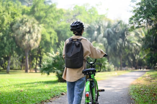 Rear view of young woman walking with bicycle in a sunny park. Lifestyle, transportation, and ECO friendly concept.