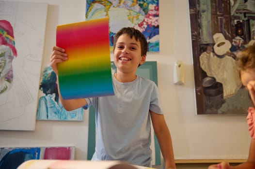 Cheerful handsome Latin American teenage boy holding a cardboard paper painted in rainbow colors, smiling looking at camera. Art class. Kids. Education