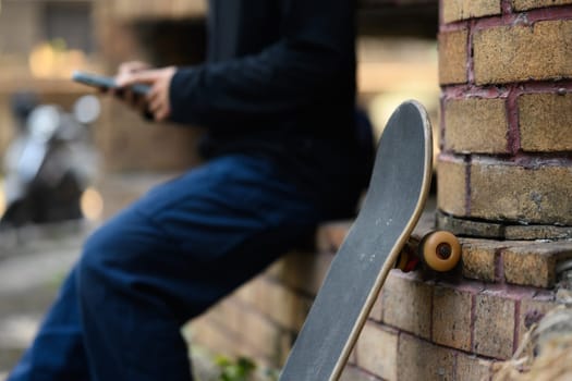 Cropped shot skateboard leaning against brick wall with man using mobile phone in background.