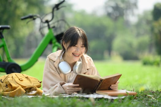 Pleased young female student reading book, lying on green grass near her bicycle in the park.