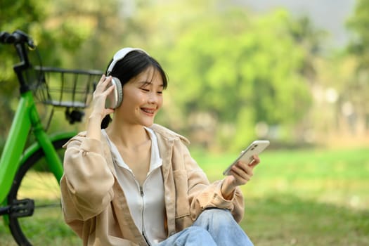 Pretty young woman listening to music with mobile phone and headphones while sitting at the park with a bicycle.