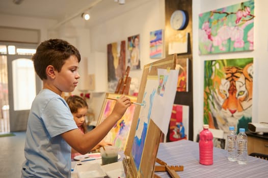 Talented primary school kid boy concentrates on painting picture on canvas in creative art classroom. Visual art. Development of artistic skills of children.