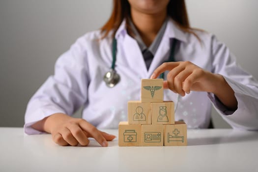 Doctor in white uniform touching wooden cubes stack in pyramid shaped with medical insurance icons.