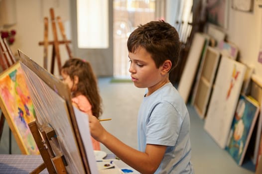 Confident portrait of a Caucasian handsome teenage school boy in blue casual shirt, holding a paintbrush and drawing picture on canvas, while learning fine art in creative art workshop