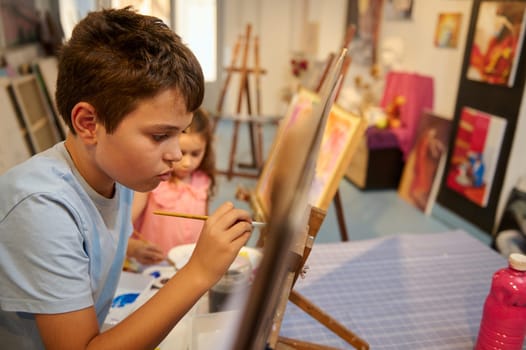 Caucasian handsome teenage school boy focused on painting on canvas in creative art workshop. Acrylic, oil or watercolor painting, drawing, people, kids, education concept