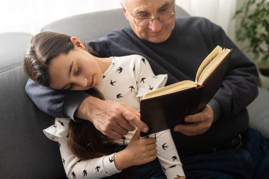 Photo of aged old grandpa little pretty granddaughter sit sofa hugging stay house quarantine safety read interesting book fairy tale modern interior living room indoors