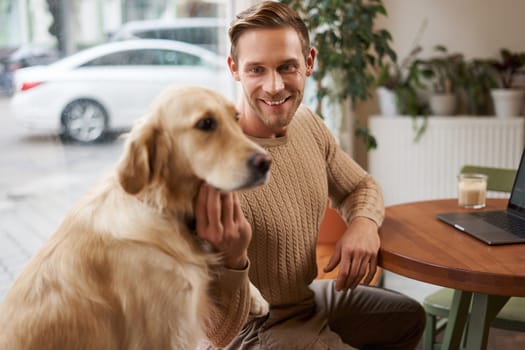 Close up portrait of smiling handsome european man with his dog in a cafe. Guy pets his golden retriever while working outdoors in coffee shop.