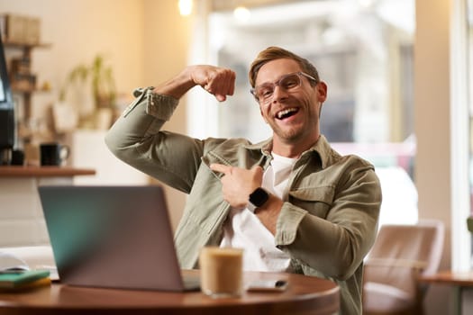 Portrait of handsome, pleased young man shows biceps, flexing muscles, sits in front of laptop in cafe, rejoicing, private coach having a video chat with his client.