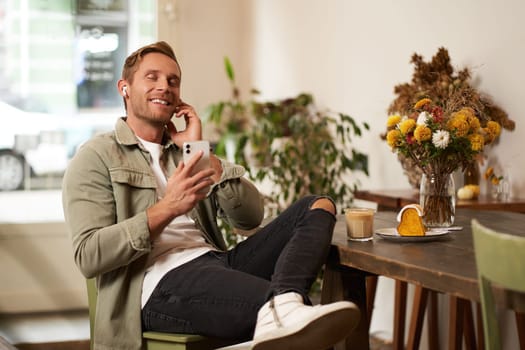 Happy handsome man in coffee shop, listening to music in wireless earphones, connects to cafe wifi to enjoy favourite song on his smartphone playlist, sitting and drinking cappuccino.