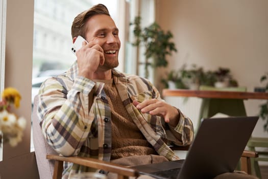 Lifestyle shot of young happy man sitting in cafe with laptop, answer phone call, talking to friend on mobile and laughing, chatting to someone with joyful face, resting in chair.