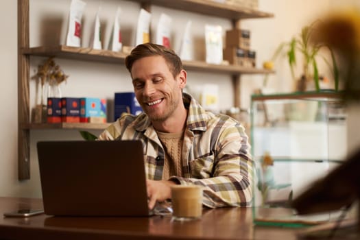 Portrait of handsome young man using laptop in cafe, sitting with computer in creative public space, typing on keyboard with pleased happy smile.