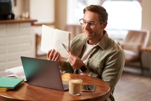 Portrait of handsome adult man showing his exercise to tutor, studying online from coffee shop, sitting in cafe with laptop, pointing at sentence in notebook.