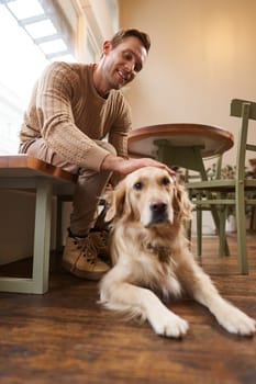 Vertical portrait of smiling handsome man with his dog, drinks coffee in pet-friendly cafe in city, touches golden retriever.