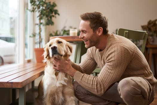 Portrait of happy smiling handsome man, pets his golden retriever and laughs. Young guy with his dog in a cafe.