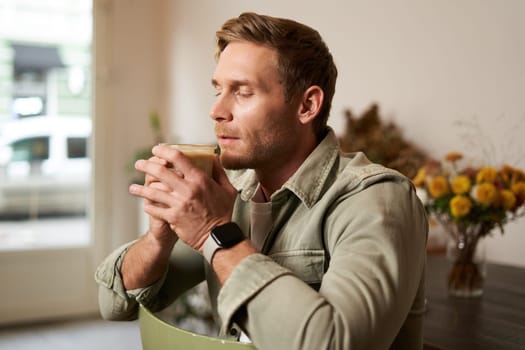 Portrait of handsome blond man in cafe, sits on chair, enjoys the taste of delicious freshly brewed cup of coffee.