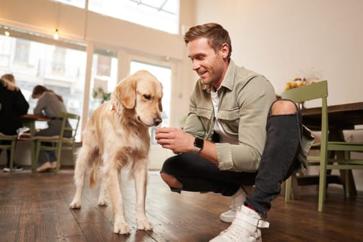 Portrait of smiling handsome man with his dog, sitting on floor in cafe with golden retriever, giving a treat.