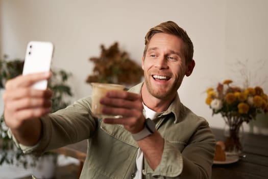 Cheerful handsome young man, taking selfie on mobile phone app, video chats to someone and raising glass of coffee, cheers gesture, drinks cup of cappuccino and smiles at smartphone camera.