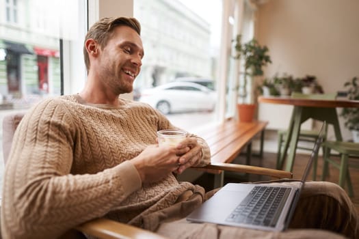 Portrait of handsome young man sits in cafe, drinks coffee and watches video on laptop, looking at screen with happy smile, relaxing in co-working space.