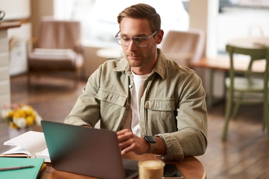 Remote work and freelance concept. Handsome man in glasses sits with notebook and laptop, studying online, working remotely from coffee shop, sitting in cafe with computer.