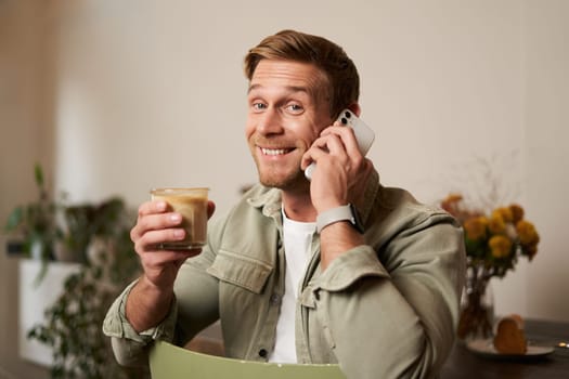 Portrait of handsome young man talking on the phone, sitting in cafe and drinking coffee, answering a call.