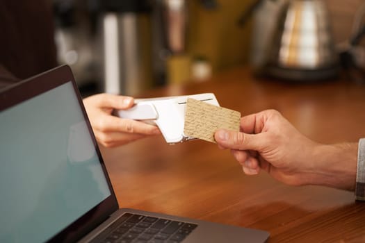 Close up cropped shot, male hand holding credit card, paying contactless by pressing it to the POS terminal, making purchase, completing order in cafe.