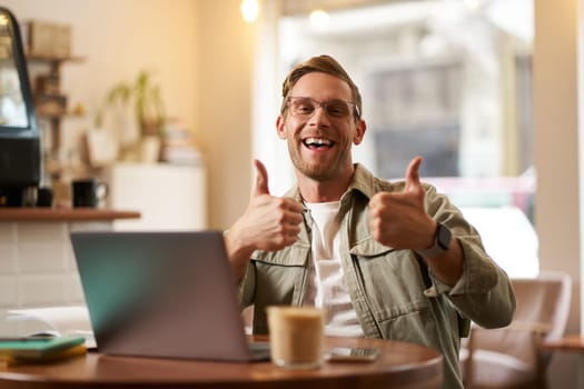 Portrait of handsome smiling man, shows thumbs up, sits in cafe with cup of coffee and laptop, approves smth good, recommends co-working space or online website.