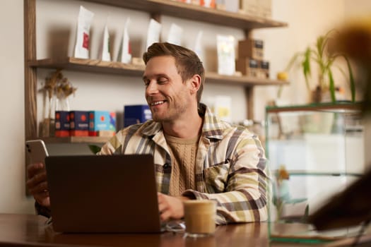 Lifestyle portrait of young businessman, freelancer sitting in cafe, drinking coffee and using laptop, working on online project, digital nomad doing his task, has remote job, looking satisfied.