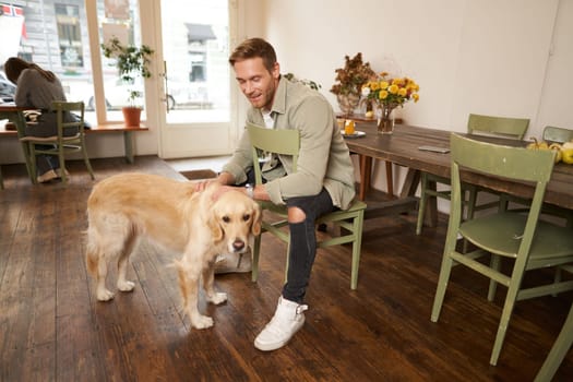 Lifestyle portrait of handsome smiling man sitting in pet friendly cafe with his beautiful dog, petting golden retriever, waiting for an order.