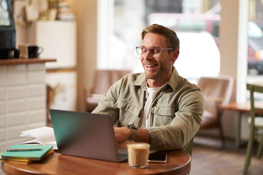 Image of young digital nomad, man in glasses sits in cafe, works from coffee shop, uses laptop in co-working space, wears glasses, drinks his beverage.