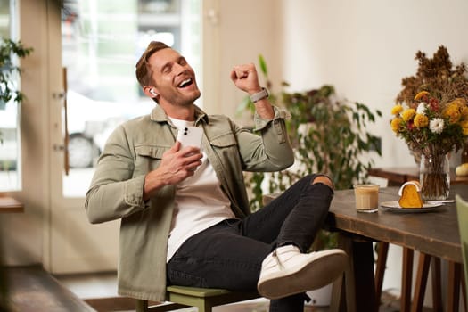 Portrait of handsome happy man in cafe, listens to music in wireless earphones, holding smartphone, connects to public wifi and enjoys favourite song, relaxing in coffee shop.