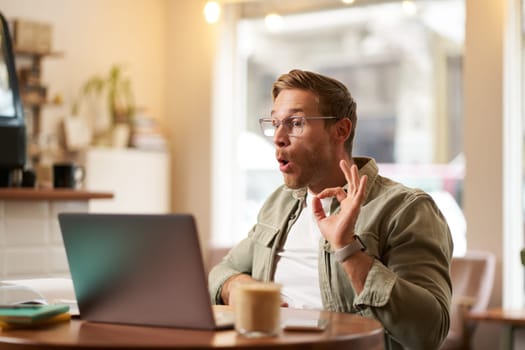 Portrait of young freelancer, digital nomad having an online meeting, video chats, shows okay sign at laptop, confirm something, makes ok hand gesture, sits in cafe, works on remote.