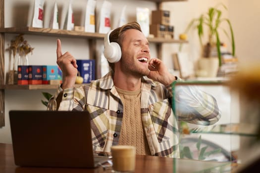 Portrait of handsome happy guy, freelancer working and listening to music in wireless headphones, dancing on his chair in cafe, enjoying favourite song in earphones.