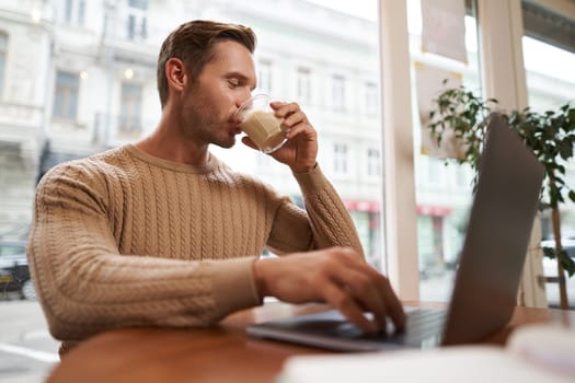 Image of handsome young businessman, man with a laptop, drinking coffee in cafe and looking at his screen, working on project in co-working space.