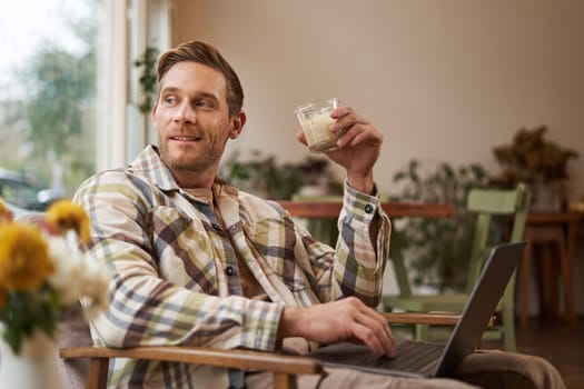 Portrait of young freelancer, a man in casual clothes, sitting in cafe and drinking coffee, working remotely on laptop, digital nomad doing his project outdoors in co-working space.