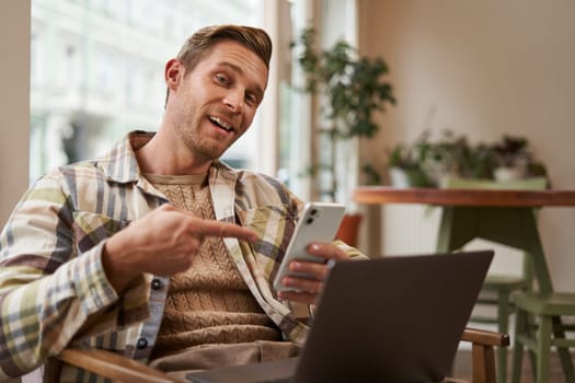 Excited guy in cafe, sitting with phone and pointing at screen with interest, recommending an app, using mobile device and laptop in co-working space. concept of freelance and remote workspace.