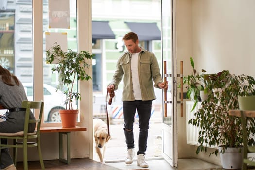 Portrait of handsome young man walks into the cafe with his dog on a leash, enters pet-friendly coffee shop, opens the door.