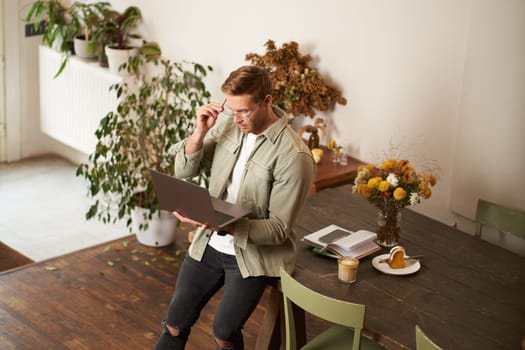 Handsome manager, man in glasses sits on table, holds laptop and types, sends message, works on project, drinks coffee, spends time at co-working space or an office.